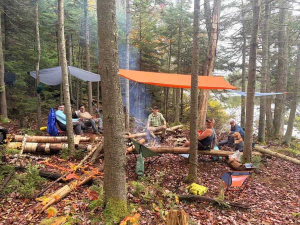 Friends sitting around a campfire under orange and gray tarps in the woods, with a dog