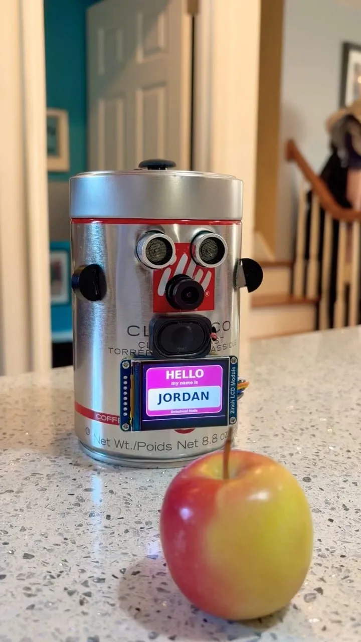 Jordan, a silver Illy coffee can with camera eyes and a small screen showing a Hello Jordan name tag, next to an apple